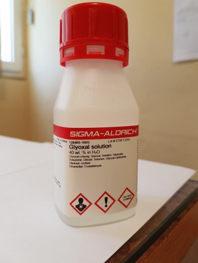 بطری محلول گلی‌اکسال (Glyoxal solution) از شرکت Sigma-Aldrich با حجم 100 گرم، درب قرمز، برچسب شامل هشدارهای شیمیایی و کد محصول 128465